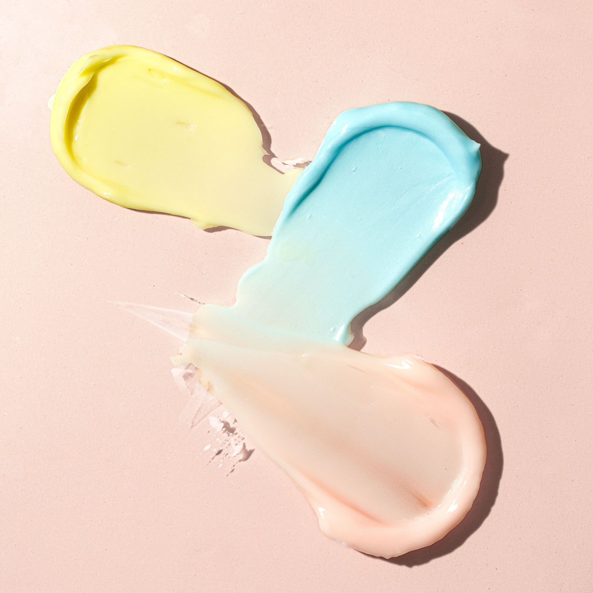 Two dollops of glossy cream, yellow and light blue, spread on a pale pink surface
