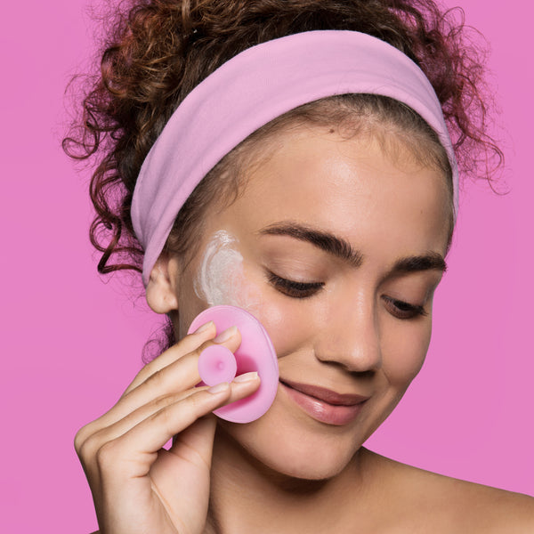 Woman using a round pink applicator pad to apply facial mask while wearing a headband